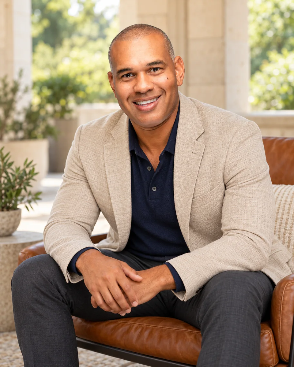 Derrick D’Acolatse seated in a light blazer and navy polo on a garden terrace